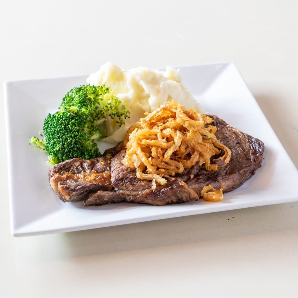 Grilled steak topped with crispy fried onions, served with mashed potatoes and steamed broccoli on a white plate.