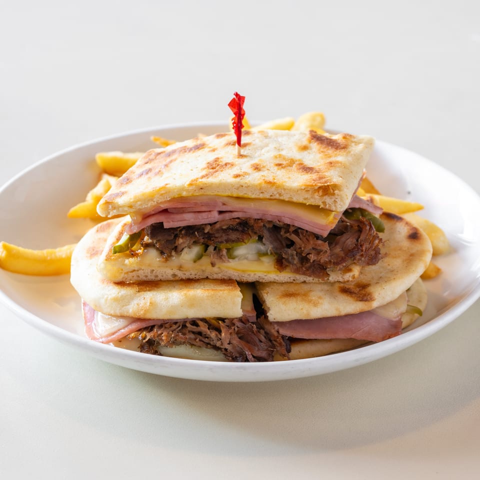 Grilled pressed sandwich layered with ham, cheese, shredded meat, vegetables, and fries on a white plate.