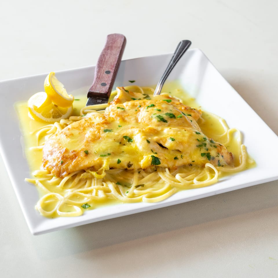Golden chicken breast with lemon cream sauce over linguine, garnished with parsley and lemon wedges on a white plate.