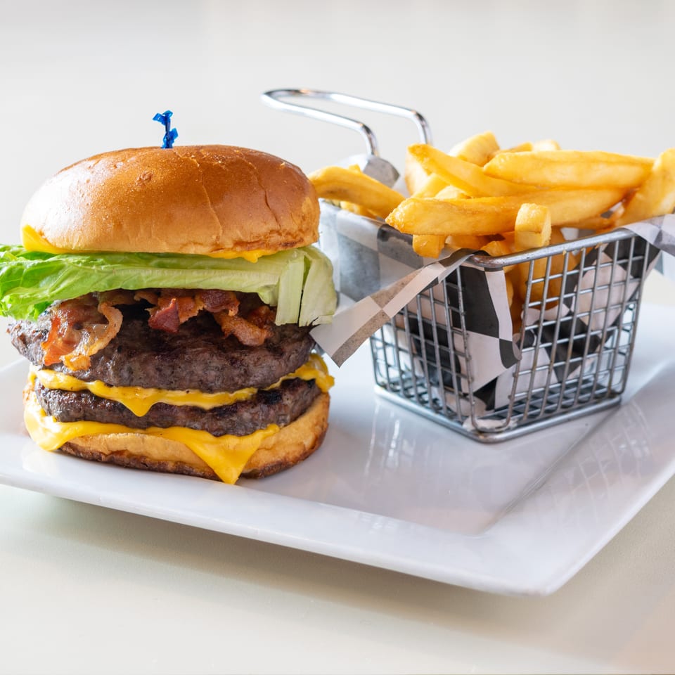 Double cheeseburger with lettuce, bacon, and melted cheese beside golden fries in a wire basket on a white plate.