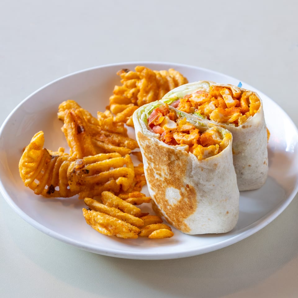Sliced fried chicken wrap with lettuce and tomatoes served alongside curly fries on a white plate.